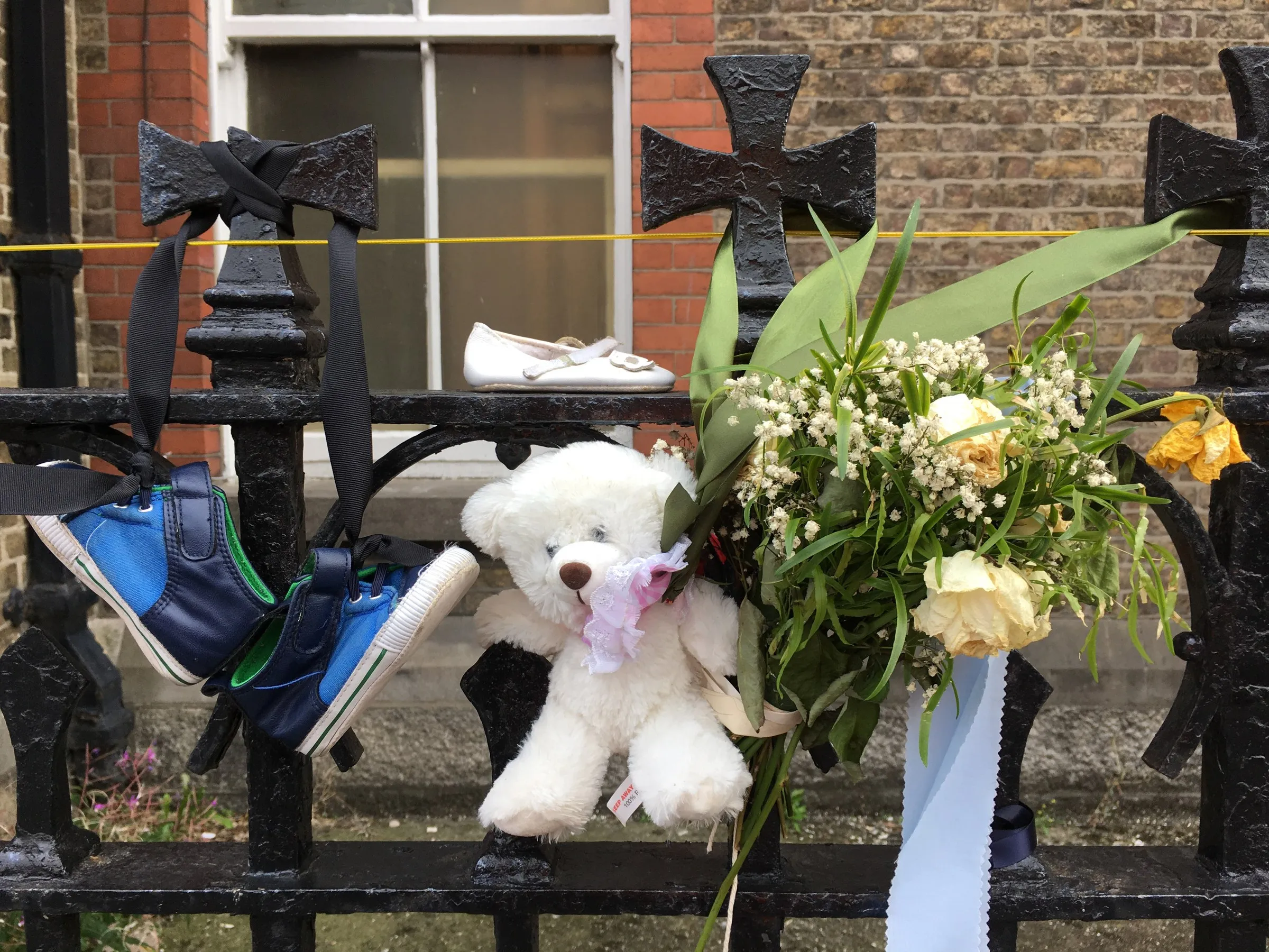 A white teddy bear, flowers and small shoes tied to iron railings