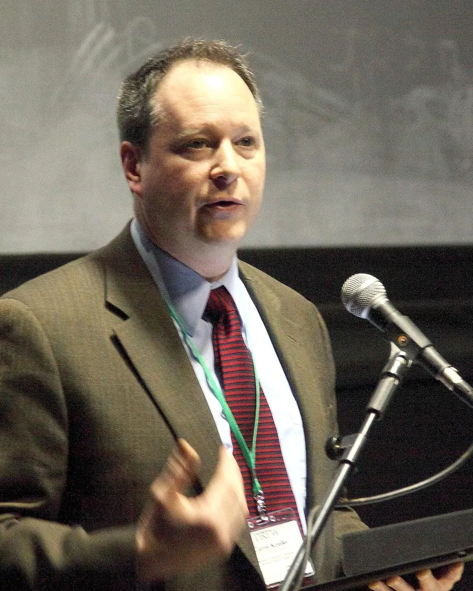 Gavin Keulks at a lectern, reading from a folder into a conference microphone
